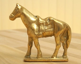 May include: A brass horse figurine with a saddle. The horse is standing on a rectangular base. The figurine is gold-colored and has detailed features, including the horse's mane, tail, and saddle.