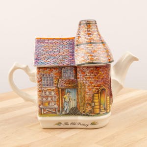 May include: A ceramic teapot shaped like a brick building with a chimney. The teapot is decorated with a colorful illustration of a pottery shop with a worker standing in the doorway. The text "The Old Pottery" is printed on the front of the teapot.