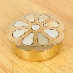 May include: A small, round, gold-colored trinket box with a flower-shaped design. The flower is made of white mother-of-pearl and has a scalloped edge.