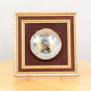 May include: A gold framed barometer with a silver face and a glass dome. The barometer has a needle that points to the current weather conditions. The frame has a brown velvet backing.