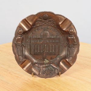 May include: A bronze ashtray with a depiction of St. Peter's Basilica in Rome. The ashtray has a scalloped edge and features two figures on either side of the basilica.