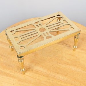 May include: A brass stove top trivet with a decorative design and four legs. The trivet is on a wooden surface.