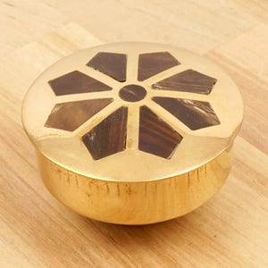 Puede incluir: Una caja redonda de latón para baratijas con una tapa decorativa. La tapa presenta un diseño de estrella en forma de estallido hecho de madera marrón oscura.