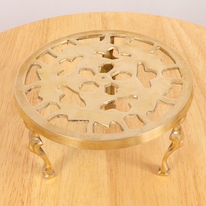 Puede incluir: Un salvamanteles de latón con un diseño decorativo calado y tres patas. El salvamanteles es redondo y tiene un patrón floral.