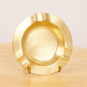 Puede incluir: Un cenicero de metal dorado con un centro redondo ligeramente hundido y un borde ondulado.