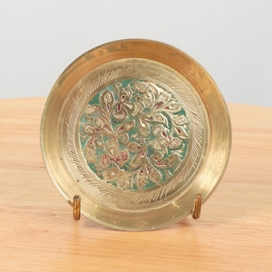 Op de afbeelding: Een klein, rond messing schoteltje met een groen en gouden bloemenmotief. Het schoteltje staat op een houten oppervlak en heeft twee kleine metalen standaards.