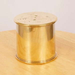 Puede incluir: Un cilindro de metal dorado con una parte superior redonda. La parte superior tiene marcas que incluyen el texto "15.24 II" y "1916".