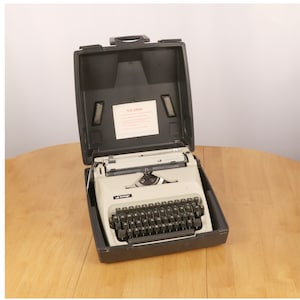 May include: A black and white typewriter with a black carrying case. The typewriter has a standard keyboard layout and a carriage return lever.
