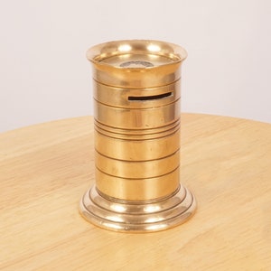 May include: A gold metal coin bank with a cylindrical shape and a slot on the side. The bank has a decorative band around the top and bottom.