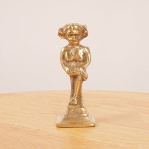 May include: A small, gold-coloured brass figurine of a seated woman with a whimsical expression. The figurine is on a square base.