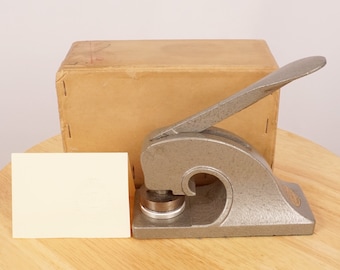 May include: A silver metal seal stamp with a handle and a round impression area. The stamp is sitting on a wooden surface next to a blank white card and a cardboard box.