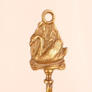 May include: A gold-toned metal key with a swan design. The swan is depicted in a nest of reeds. The key has a ring at the top for hanging.