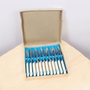 Könnte beinhalten: Ein Set Vintage-Gabeln mit weißen Griffen und silbernen Zinken, ordentlich in einer Pappschachtel mit blauem Innenraum angeordnet. Die Gabeln sind wahrscheinlich aus Edelstahl.