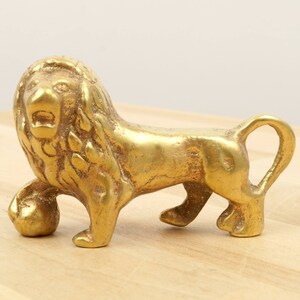 May include: A small, gold-colored brass lion figurine with a ball in its paw. The lion is standing on a wooden surface.