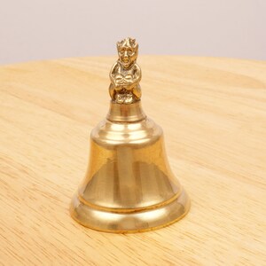 May include: A gold-colored bell with a small, detailed figurine of a creature with a crown on top. The bell is sitting on a wooden surface.