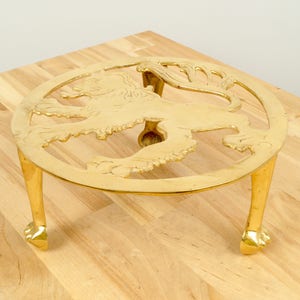 Puede incluir: Un salvamanteles de metal dorado con un diseño de león. El salvamanteles tiene tres patas y es redondo.