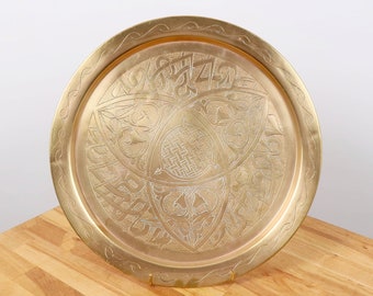 May include: A round, brass serving tray with an intricate, engraved design. The design features a central motif of a geometric pattern surrounded by floral and scrollwork.