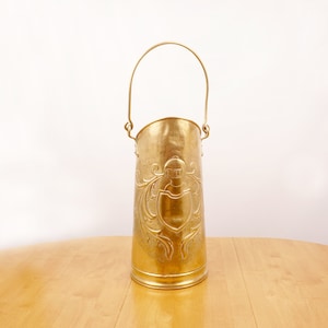 Puede incluir: Un cubo de metal dorado con asa y un diseño en relieve. El cubo está sobre una mesa de madera.