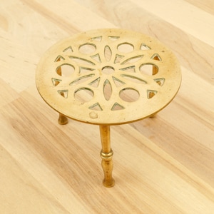 Puede incluir: Un salvamanteles de metal dorado con una parte superior circular y tres patas. La parte superior tiene un diseño decorativo calado con un patrón circular.