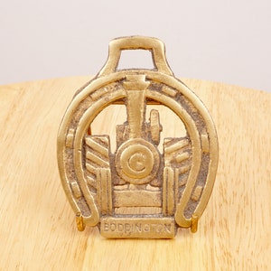 May include: A gold-colored metal horseshoe-shaped doorstop with a tractor design and the word "Boddington" engraved on the bottom.