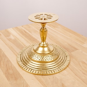 May include: A gold metal decorative stand with a round top and three legs. The top has a circular design with a small hole in the centre. The base of the stand has a decorative pattern of triangles.