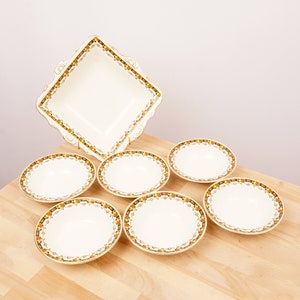 May include: A set of vintage cream-colored dishes with a decorative orange, brown, and gold border. The set includes six small bowls and one square serving dish with scalloped edges. The dishes are arranged on a light wood surface.