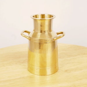 Puede incluir: Un jarrón de metal dorado en forma de bidón de leche con dos asas. El jarrón está sobre una superficie de madera.