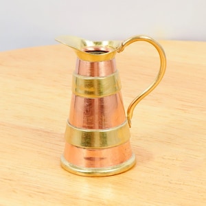 May include: A small copper and brass jug with a flared lip and a handle. The jug has a banded design with alternating copper and brass bands.