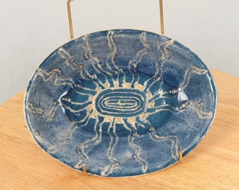 May include: A blue ceramic oval dish with a white sunburst design. The dish has a gold metal stand.