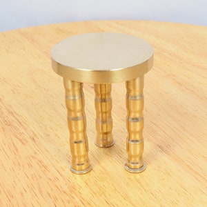 May include: A small, gold-colored metal stand with a round top and three legs. The stand is made of brass and has a simple, modern design.