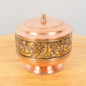 May include: A decorative jar made of copper and brass with a lid. The jar features an ornate design of fruit and leaves around the middle.