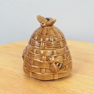May include: A ceramic honey pot shaped like a beehive with a bee on top. The pot is brown and has a textured surface.