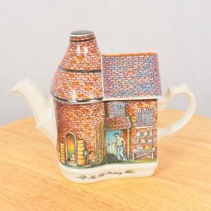 May include: A ceramic teapot shaped like a brick building with a chimney. The teapot is decorated with a colorful illustration of a pottery shop with a man standing in the doorway. The text "The Old Pottery" is printed on the bottom of the teapot.