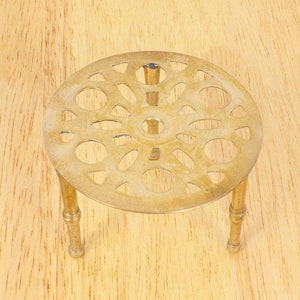 Könnte beinhalten: Ein Messing-Topfuntersetzer mit einem dekorativen, runden Design und drei Beinen. Der Topfuntersetzer befindet sich auf einer Holzoberfläche.