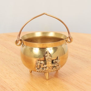 Könnte beinhalten: Ein kleiner, messingfarbener Kessel mit gebogenem Griff und drei kleinen Beinen. Der Kessel zeigt ein Relief von Cardiff Castle mit dem Text "Cardiff Castle" darunter. Der Griff hat eine hellere Messingfarbe.