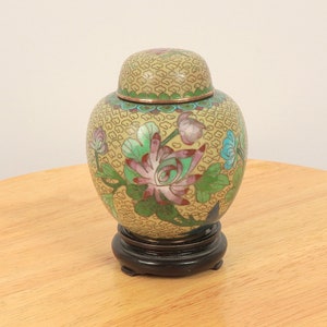 Op de afbeelding: Een kleine, ronde, gele cloisonné pot met een bloemenmotief. De pot heeft een deksel en staat op een houten voet.