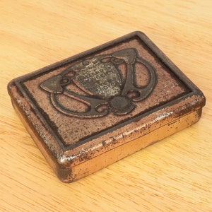 May include: A vintage, rectangular, metal tin box with a tarnished copper finish. The box features an ornate, embossed design with a crest and the text "Gillard & Bowyer's, Bristol, Eng."
