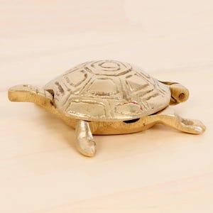 May include: A brass turtle figurine with a hinged shell. The turtle has detailed markings on its shell and is resting on a light-colored surface. The figurine is a decorative item.