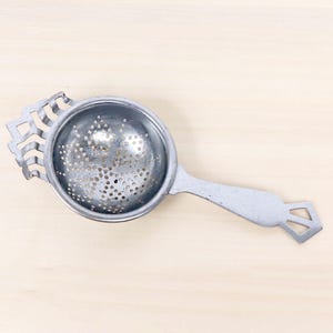 May include: A silver-toned metal tea strainer featuring a circular bowl with a perforated mesh surface, a decorative stepped handle end, and a notched, tiered hook on the opposite side for resting the tool on the rim of a cup or mug.