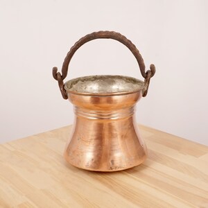 Può includere: Una pentola di rame con finitura martellata e manico in ferro. La pentola ha un fondo arrotondato e un bordo svasato.