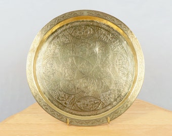 May include: A round, gold-colored metal tray with an intricate, embossed design. The tray has a raised rim and a smooth, polished surface.