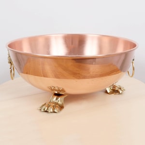 May include: A copper bowl with a polished finish, featuring brass lion's paw feet and ring handles. The bowl has a wide, open design and a reflective surface, ideal for decorative or functional use. The bowl is approximately 30 cm in diameter.