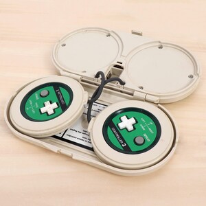 May include: A vintage video game controller with two circular control pads. Each pad has a green center with a white cross-shaped directional pad, a gray button, and the word "SPRAY". The controller is beige and connected by a gray cable.
