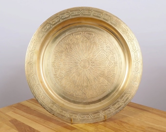 May include: A round, gold-colored metal serving tray with an intricate, embossed design. The tray has a raised rim and a decorative border.