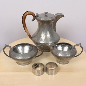 Pode incluir: Um conjunto de chá de peltre vintage com acabamento martelado. O conjunto inclui um bule com alça de madeira, um creamer, um açucareiro e dois anéis de guardanapo.