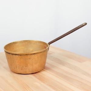 May include: A vintage brass pot with a long iron handle. The pot is sitting on a wooden surface.