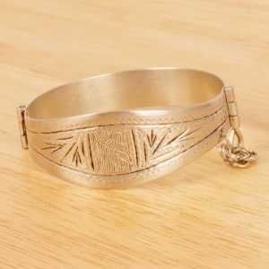 Könnte beinhalten: Ein silbernes Armband mit einem komplizierten gravierten Design. Das Armband hat einen Scharnierverschluss und eine Kette, die am Ende befestigt ist.