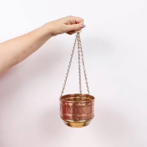 Pode incluir: Um vaso suspenso com um corpo cilíndrico de cobre com um padrão de grade texturizado e uma base de latão lisa. O vaso é suspenso por três correntes de metal finas que se encontram num anel circular no topo para pendurar.