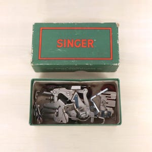 May include: A green rectangular box with the red text SINGER printed on the lid. The box is open, revealing a collection of various silver-coloured metal sewing machine attachments and presser feet stored inside the cardboard container.