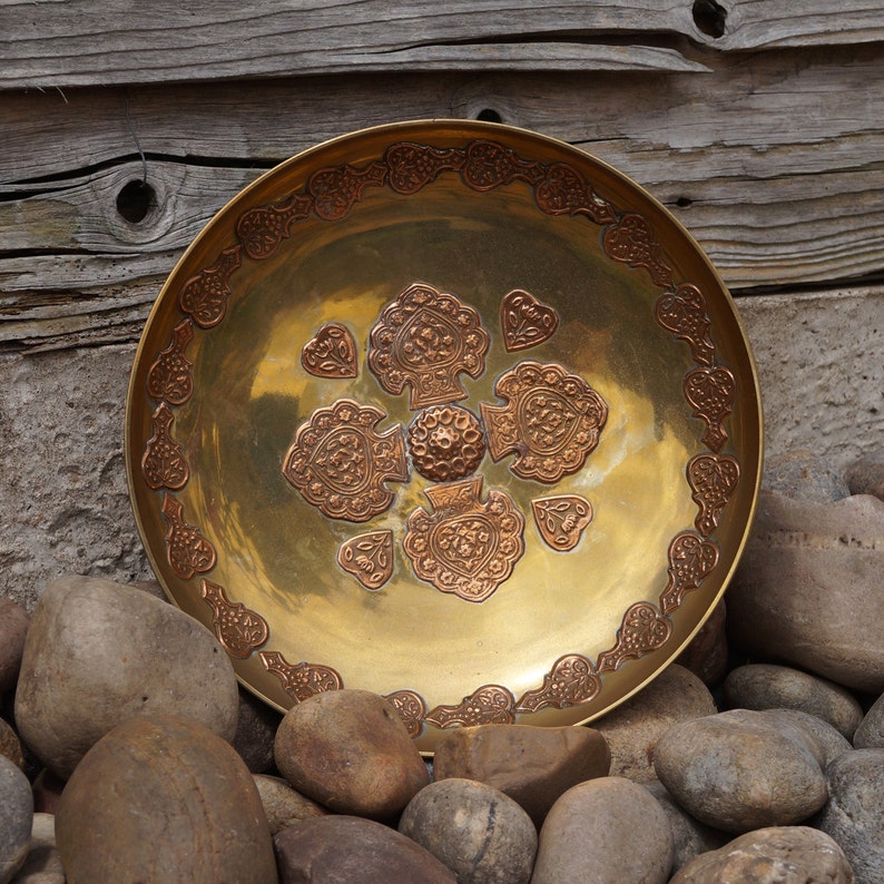 May include: A round, brass serving dish with an intricate, embossed design of hearts and swirls. The dish has a gold finish and is surrounded by small, brown stones.